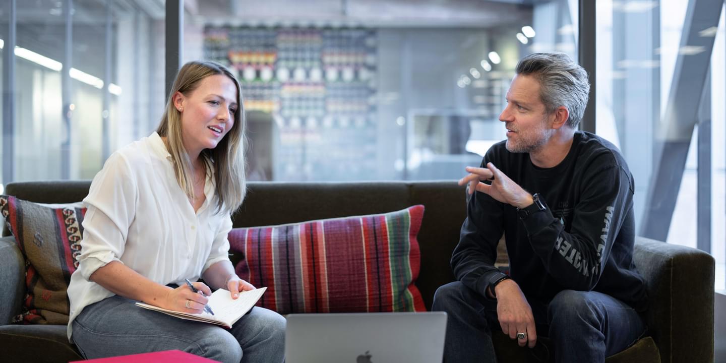 A white female and an older white male discuss a work-related topic while seated in a modern office setting with comfortable furniture. A laptop is on the coffee table in front of the couch they're sitting on. Photo by LinkedIn Sales Solutions via Unsplash.