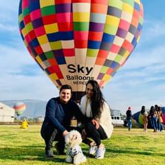 Image of Linda Villarreall, her husband, and her dog. They are posing outside on a sunny day in front of a colorful hot air balloon.