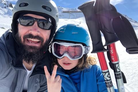 Propeller employee Noel Coffman poses on a snowy mountain with ski poles with his young child. They are both wearing helmets and eye protection. Profile Photo