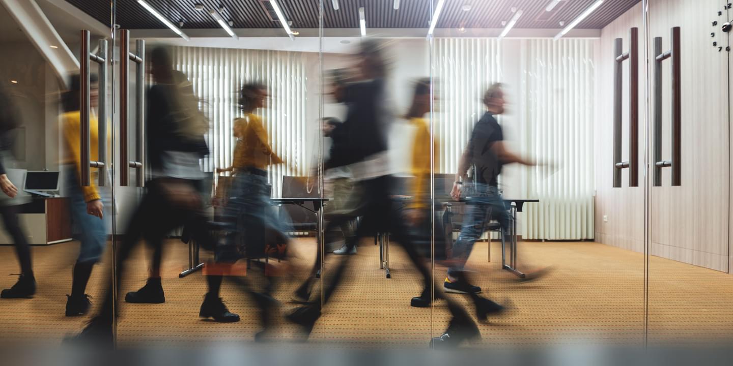 Several people walking with a blurred motion in an office looking through glass doors windows and sunlight shining through.