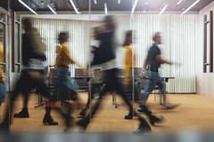 Several people walking with a blurred motion in an office looking through glass doors windows and sunlight shining through.