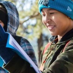 Three children dressed warmly in beanies, gloves, and jackets, consult their Outdoor Lab Foundation guide book during a nature hike. They are smiling and happy.