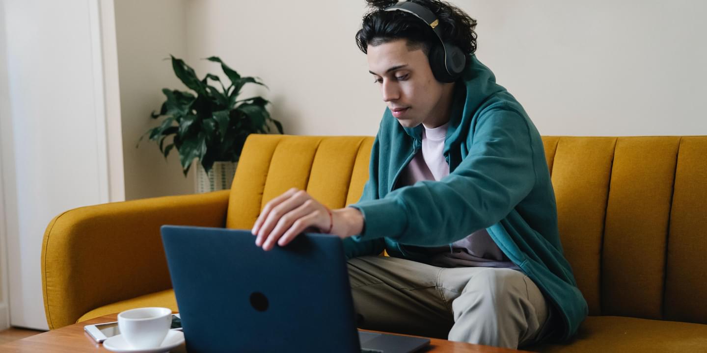 Serious man of color in wireless headphones using a laptop on a coffee table in front of a couch. It appears he is working remotely. Photo by Eren Li via Unsplash.
