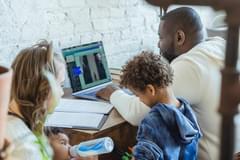 A image of a Black father working on a laptop from home while his wife feed their infant child, and their young child stands near his father's desk. Photo by Keira Burton via Unsplash.