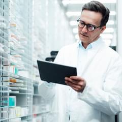 An older, white male pharmacist consults a digital tablet while surrounded by cases with dozens of shelves of medications.
