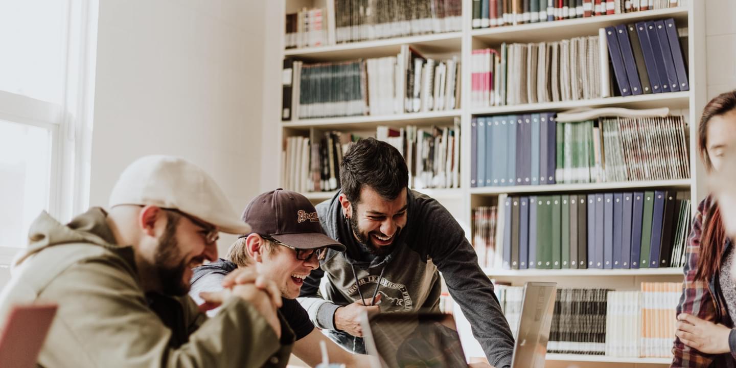 A group of young, male office employees in casual attire laugh and smile while working together on a project in a modern, bright collaboration space. Photo by Priscilla Du Preez via Unsplash.