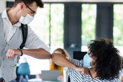 Two coworkers in an office, a white man and a Black man, bump elbows while wearing face masks during the COVID-19 pandemic.