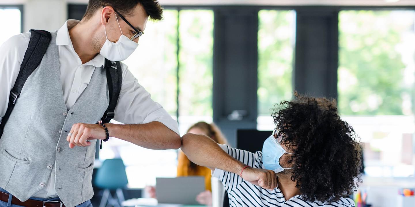 Two coworkers in an office, a white man and a Black man, bump elbows while wearing face masks during the COVID-19 pandemic.