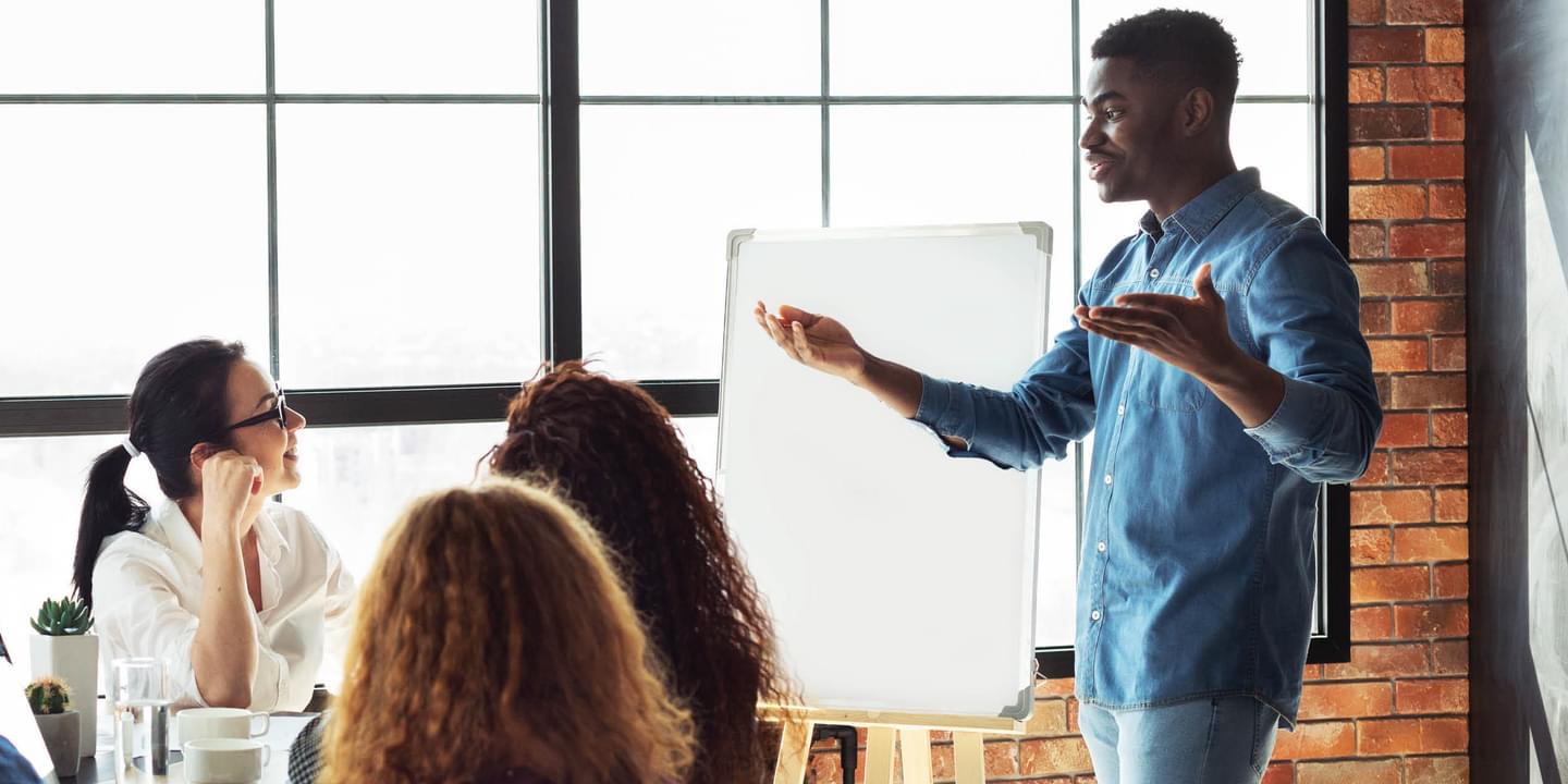 Seven business people are gathered around a meeting table. They are all of various ages, races, and genders. A Black man is standing and presenting to the rest of the group.