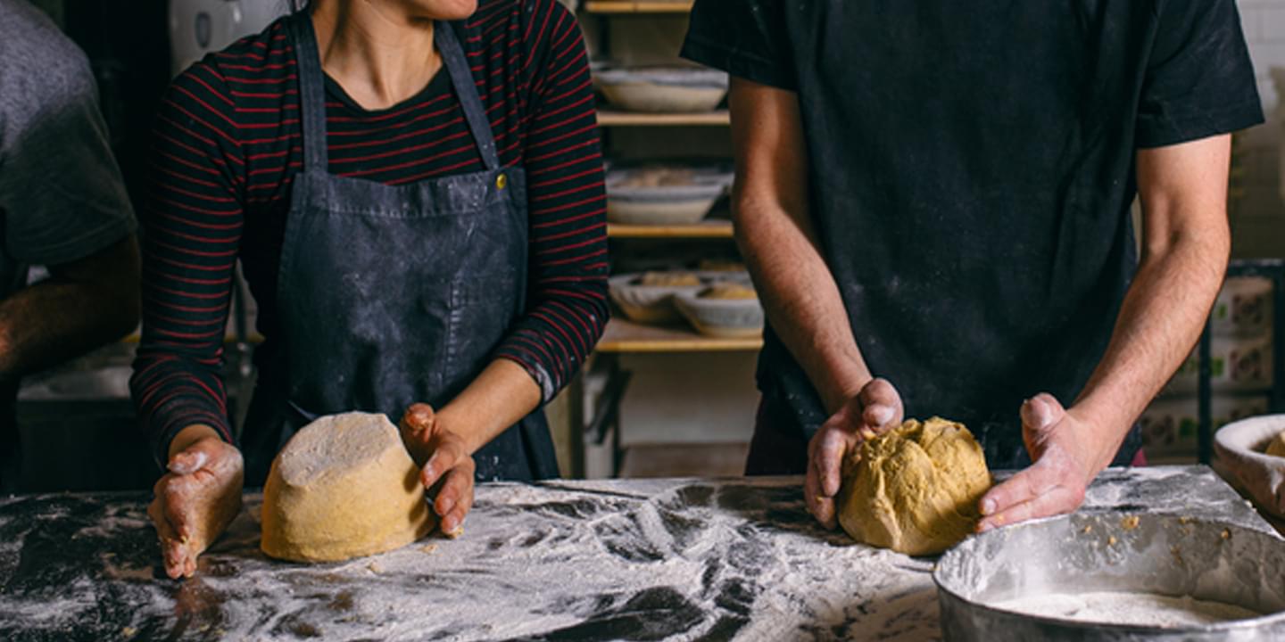 Two people knead dough on a well-floured surface in a commercial kitchen.