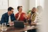 Group of four people at a meeting table discuss their project. They are gathered around a laptop on the table.