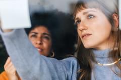 Two businesswomen brainstorming using sticky notes in an office with the viewpoint looking through the window at them