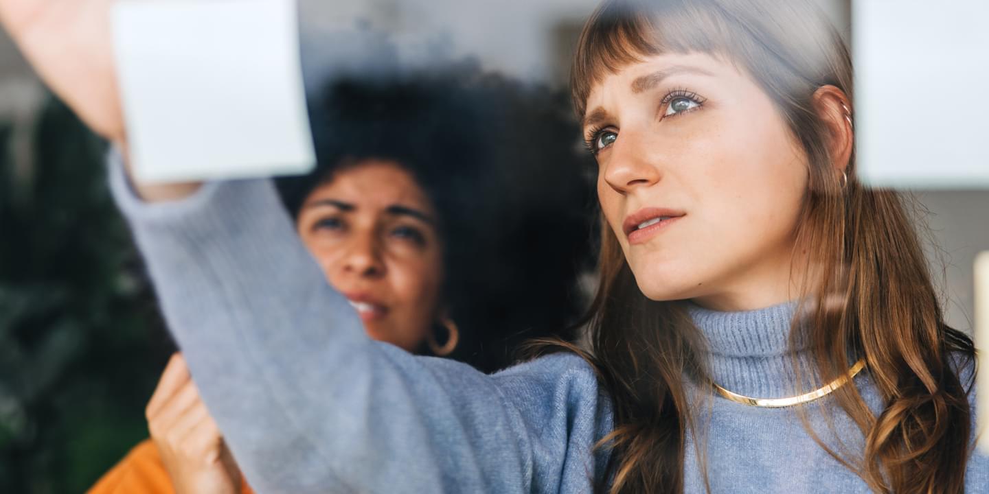 Two businesswomen brainstorming using sticky notes in an office with the viewpoint looking through the window at them