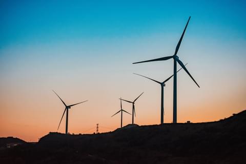Silhouettes of energy windmills at dawn – the sky is blue at the top of the image and shifts to a pale orange at the bottom of the image near the horizon. Photo by Zhang Fengsheng via Unsplash.
