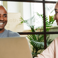 A Black man and an Asian woman collaborate sitting side-by-side with laptops. They are smiling and laughing.