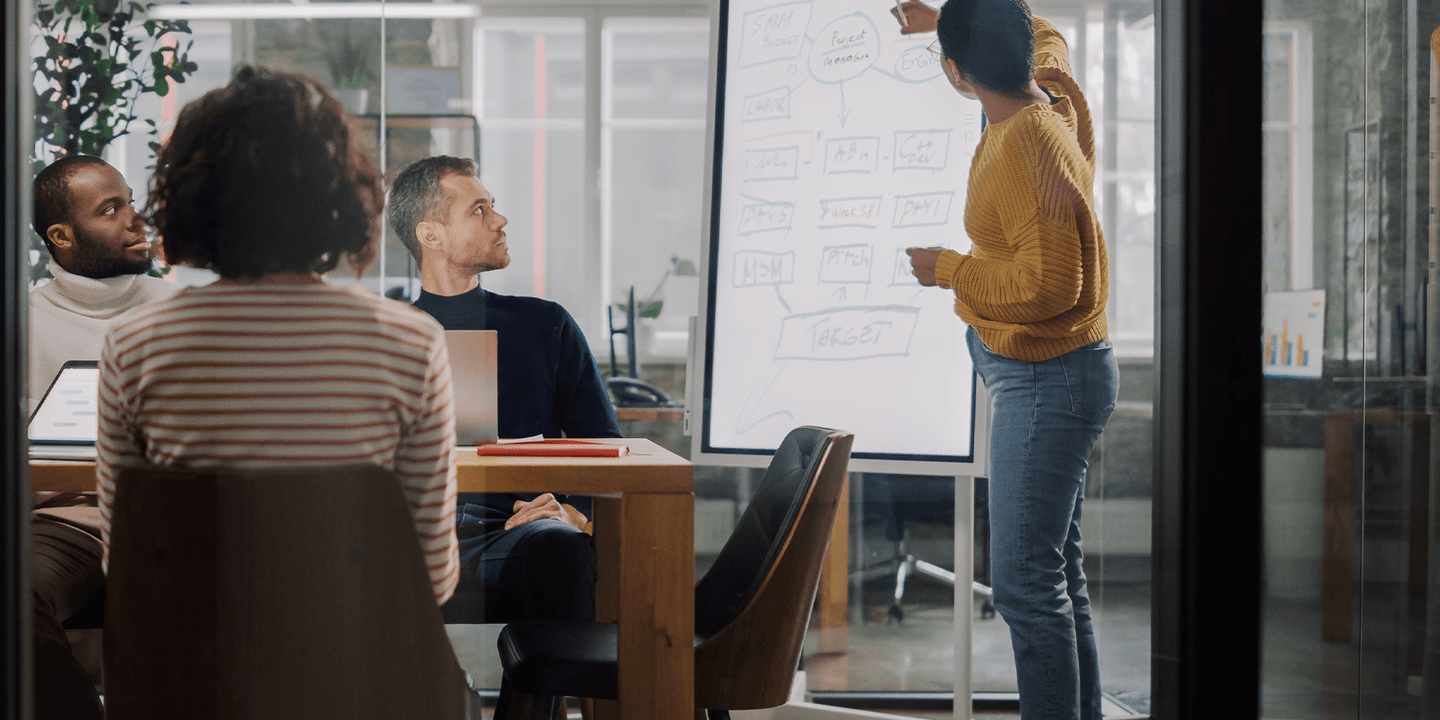 Business people of various ages, races, and genders meet in a meeting room with glass walls. One business person writes on a whiteboard while the other people watch. The image was photographed from the outside of the glass walls.