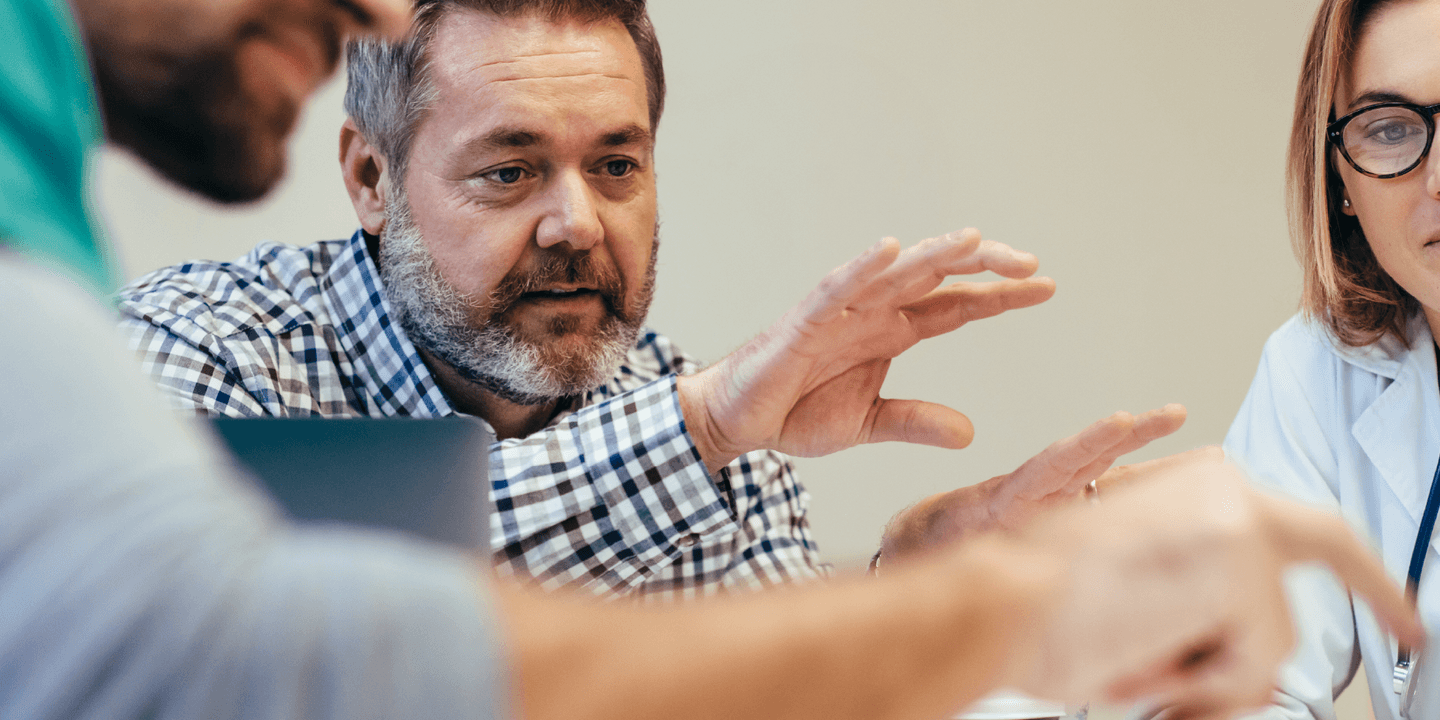 Three medical professionals gather around a digital tablet and discuss the items on the tablet's screen during a meeting.