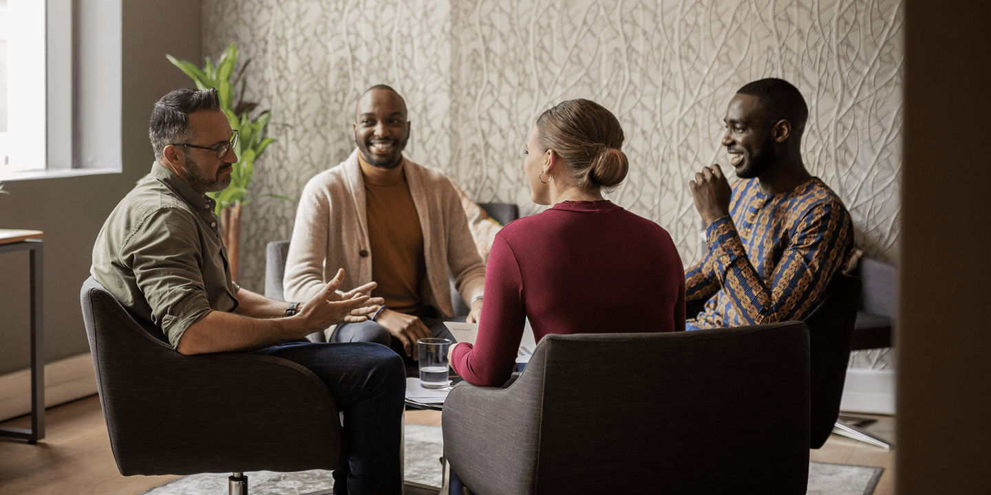 A group of four business people of various ages, races, and genders gather around a coffee table and engage in a lively conversation.