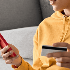 Close up of a person wearing a yellow hoodie holding a credit card in one hand and their smart phone in the other hand, implying they're making an online purchase through their mobile phone.