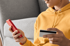 Close up of a person wearing a yellow hoodie holding a credit card in one hand and their smart phone in the other hand, implying they're making an online purchase through their mobile phone.