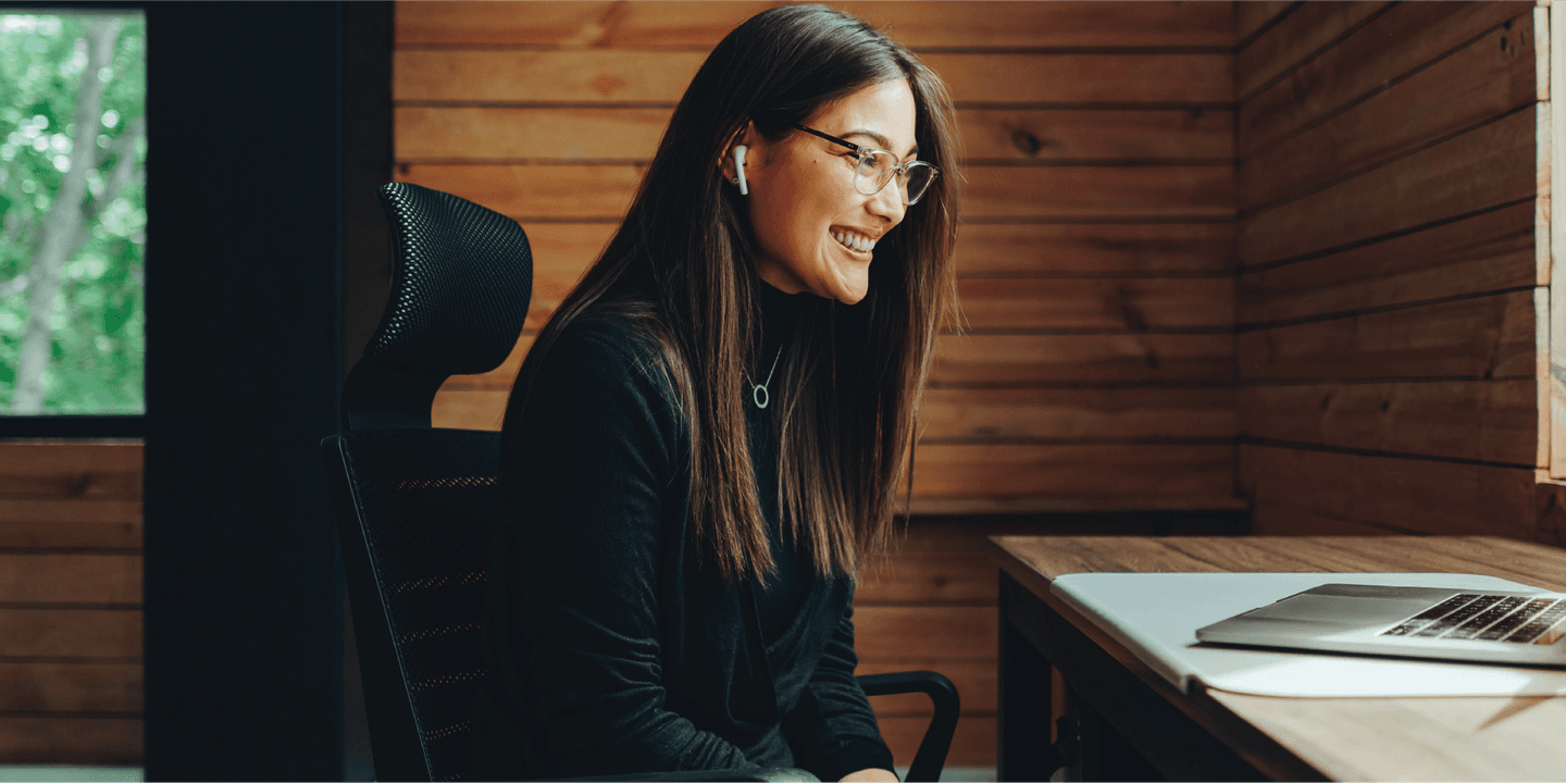 Belonging in a Hybrid World A Woman Smiling at a laptop while sitting at a desk in a room with wooden walls.