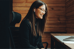Belonging in a Hybrid World A Woman Smiling at a laptop while sitting at a desk in a room with wooden walls.