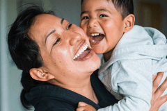 Woman of color holds up a young child of color – the child appears to be between 3 and 5 years old. They are both laughing and smiling.