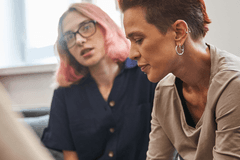 Two queer people solemnly share a moment. The person in the background seems to be comforting the person in the foreground. The shoulder of a third participant in the convo is close to the camera, but the rest of the person is located off-screen.