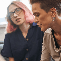 Two queer people solemnly share a moment. The person in the background seems to be comforting the person in the foreground. The shoulder of a third participant in the convo is close to the camera, but the rest of the person is located off-screen.