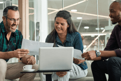 Image of three business people: the left-most is an older white man, the middle is a woman of color, the right-most is a Black man. They are gathered around a small coffee table with a laptop on it. The white man and Black man are comparing printed pages while the woman of color joins the conversation.