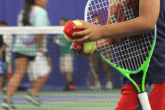 Close up of a child of color holding a tennis racket under their arm and carrying tennis balls in both hands.