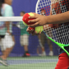 Close up of a child of color holding a tennis racket under their arm and carrying tennis balls in both hands.