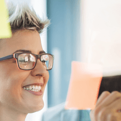 Image of business people working on a glass office partition, placing sticky notes on the glass for a project they're working on. They appear to be happy.