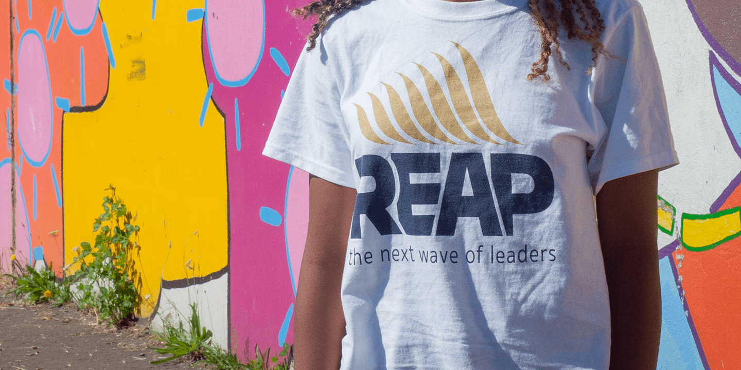 Close up of a young Black girl wearing a REAP t-shirt in front of an outdoor wall that is painted with a colorful mural.