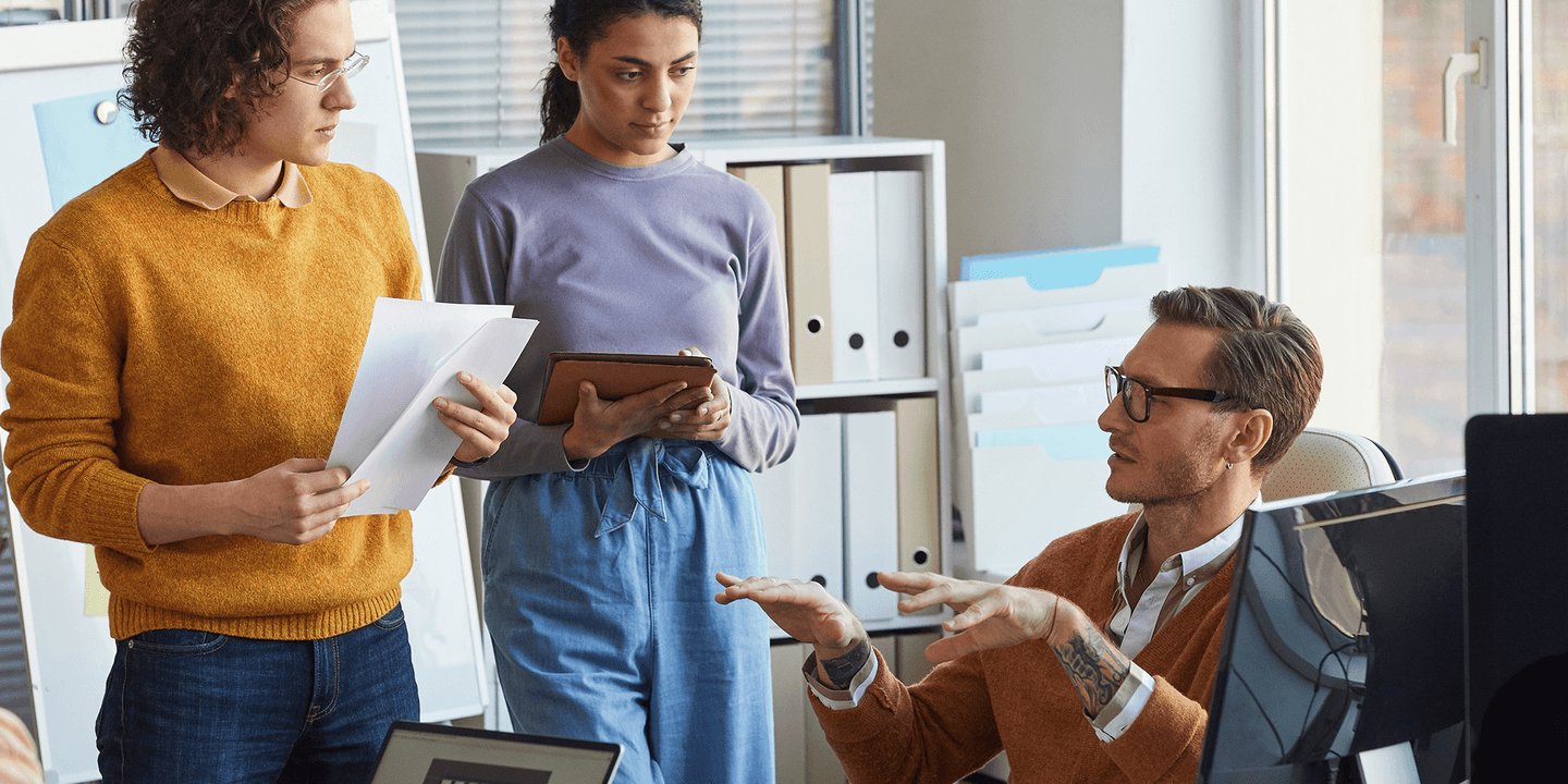 Group of business professionals of various races, ages, and genders have a discussion next to a desk with computers.