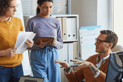 Group of business professionals of various races, ages, and genders have a discussion next to a desk with computers.