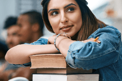 A light-skinned woman with dark hair and a denim shirt leaning on a pile of books with a slight smile on her face.